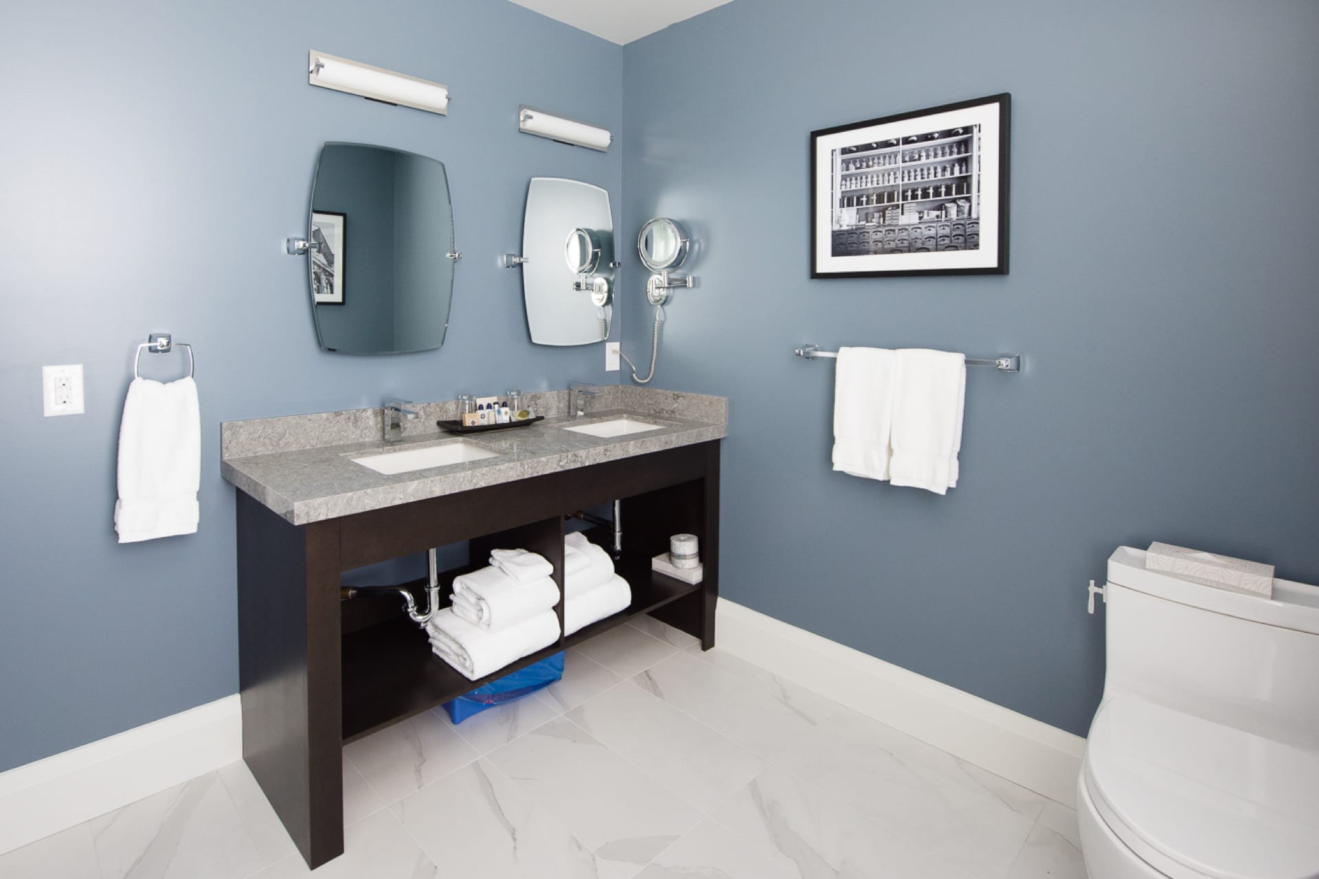 Modern bathroom with blue walls and white tile floor. Features a double sink vanity with a large mirror, round magnifying mirror, and toiletries. White towels hang beside it. A framed black and white photo is on the wall above two towel bars. 124 on Queen Hotel and Spa | Hotel & Spa Niagara-on-the-lake | 1 (855) 988-4552