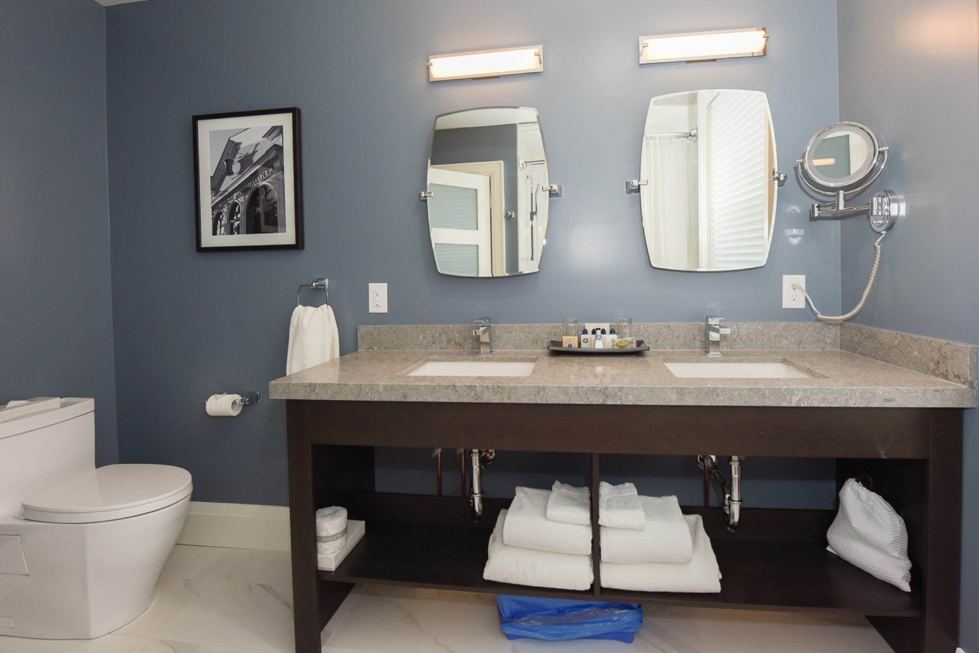 Modern bathroom with blue walls featuring a dual sink vanity with two mirrors and a light above each. A toilet is on the left, and a black-and-white photo hangs on the wall. Towels and toiletries are neatly arranged under the sinks. 124 on Queen Hotel and Spa | Hotel & Spa Niagara-on-the-lake | 1 (855) 988-4552