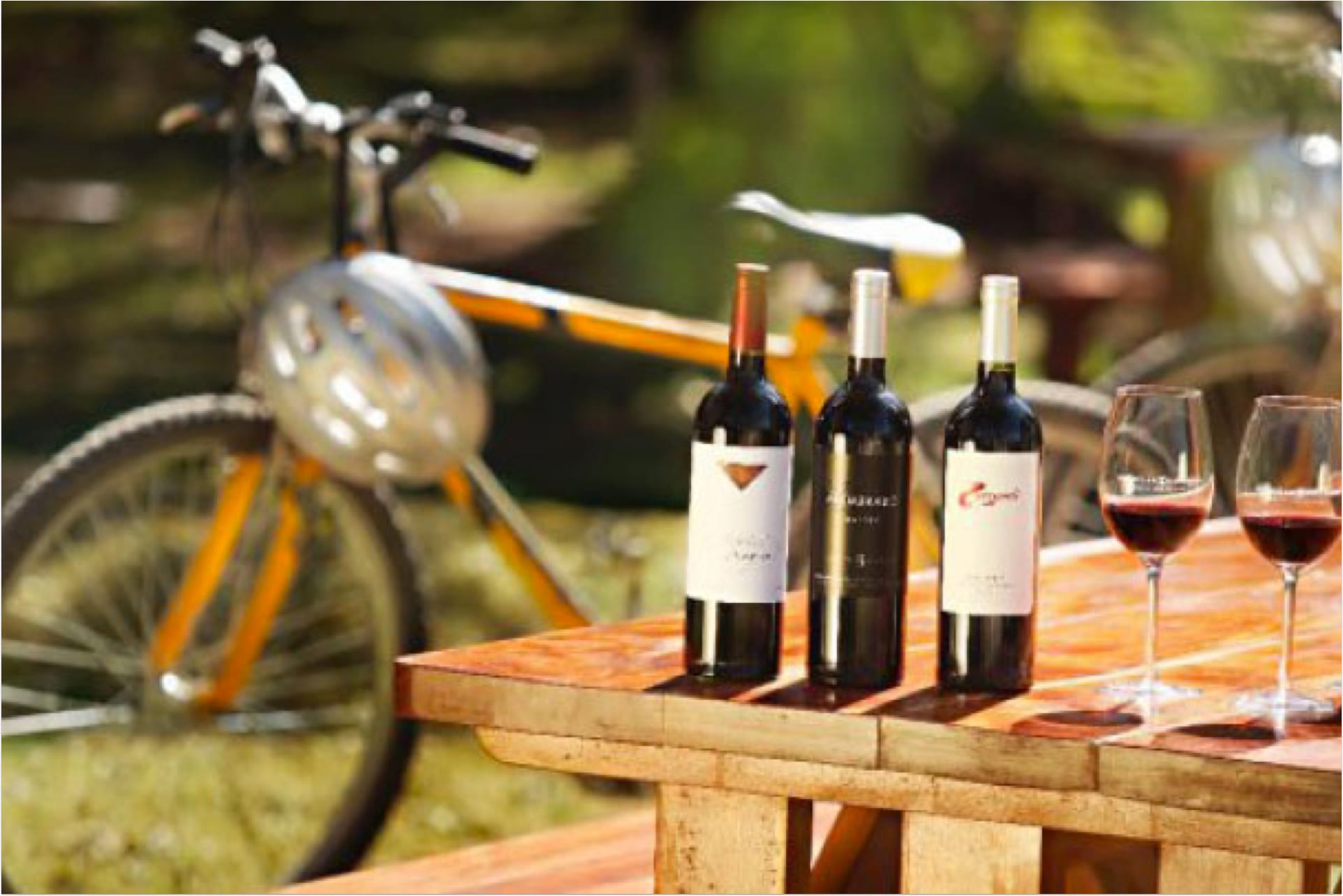 Three wine bottles and two filled glasses sit on a wooden table outdoors. A bicycle and greenery are blurred in the background, suggesting a serene setting. 124 on Queen Hotel and Spa | Hotel & Spa Niagara-on-the-lake | 1 (855) 988-4552