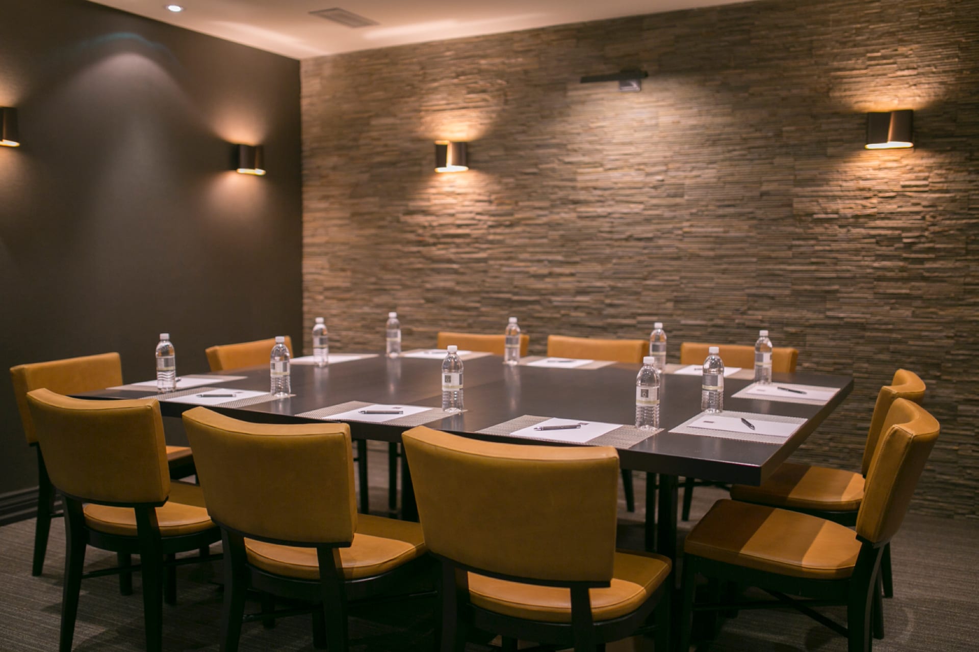 A conference room with a rectangular table set for a meeting. The table has ten chairs, each with a notepad and pencil. Bottled water is placed at each seat. The room has stone-textured walls and warm lighting. 124 on Queen Hotel and Spa | Hotel & Spa Niagara-on-the-lake | 1 (855) 988-4552