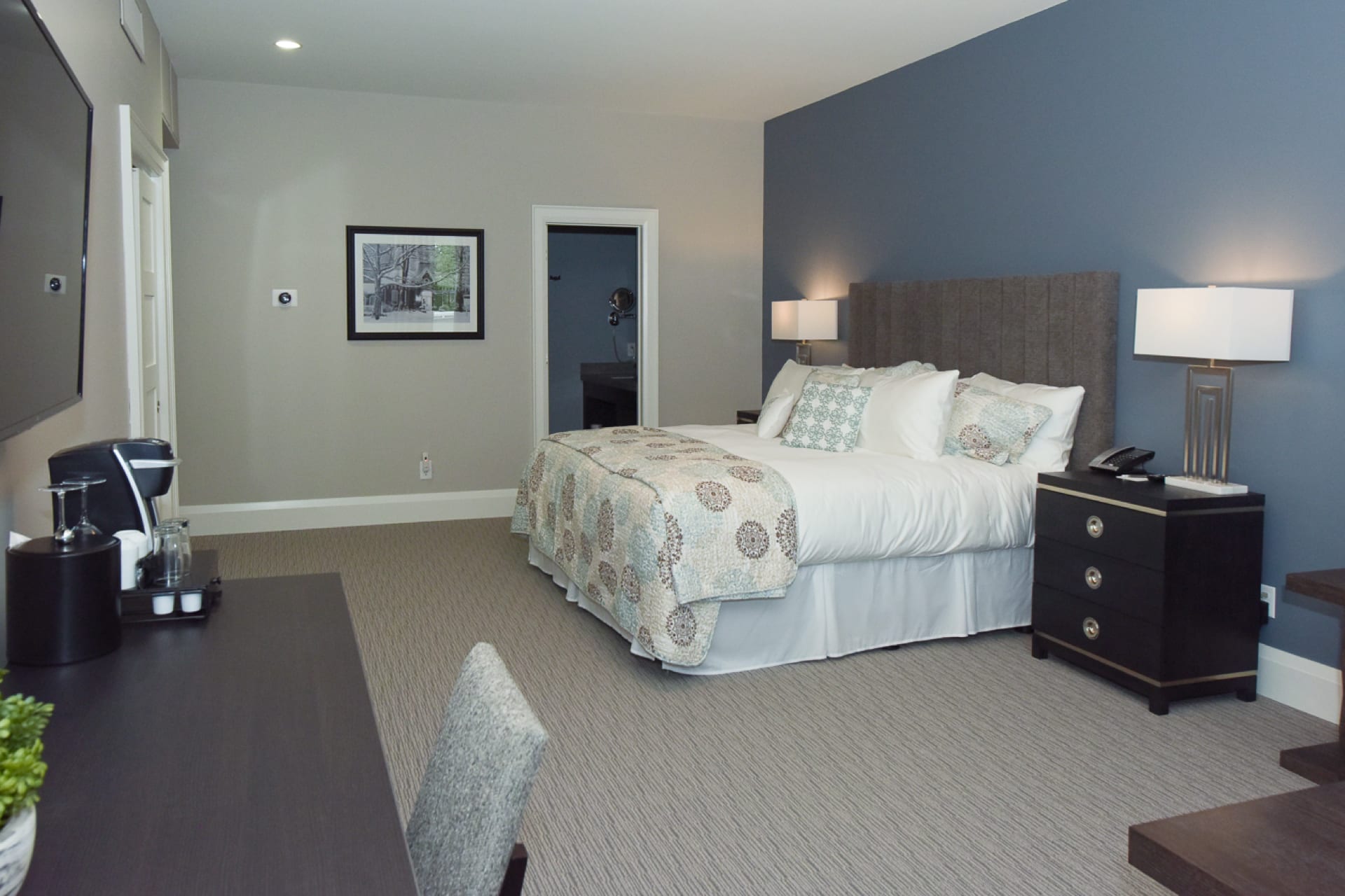 Hotel room with a neatly made bed, quilted bedding, and two bedside tables with lamps. The walls are painted blue and white. A desk with a chair and coffee maker is on the left, and a framed picture hangs above. Carpeted floor throughout. 124 on Queen Hotel and Spa | Hotel & Spa Niagara-on-the-lake | 1 (855) 988-4552