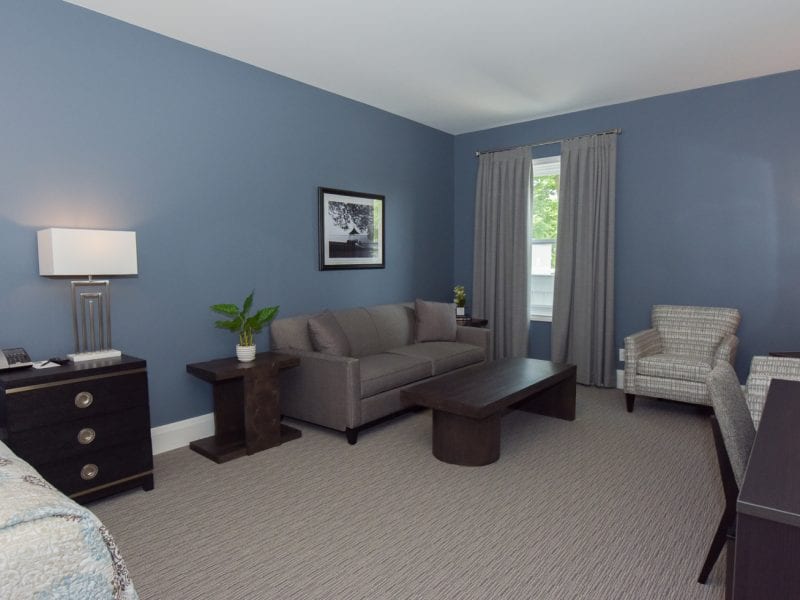 A minimalist living room with blue walls featuring a gray sofa, a wooden coffee table, two patterned armchairs, and a framed black-and-white photo. Two lamps on side tables and a window with gray curtains complete the space. 124 on Queen Hotel and Spa | Hotel & Spa Niagara-on-the-lake | 1 (855) 988-4552