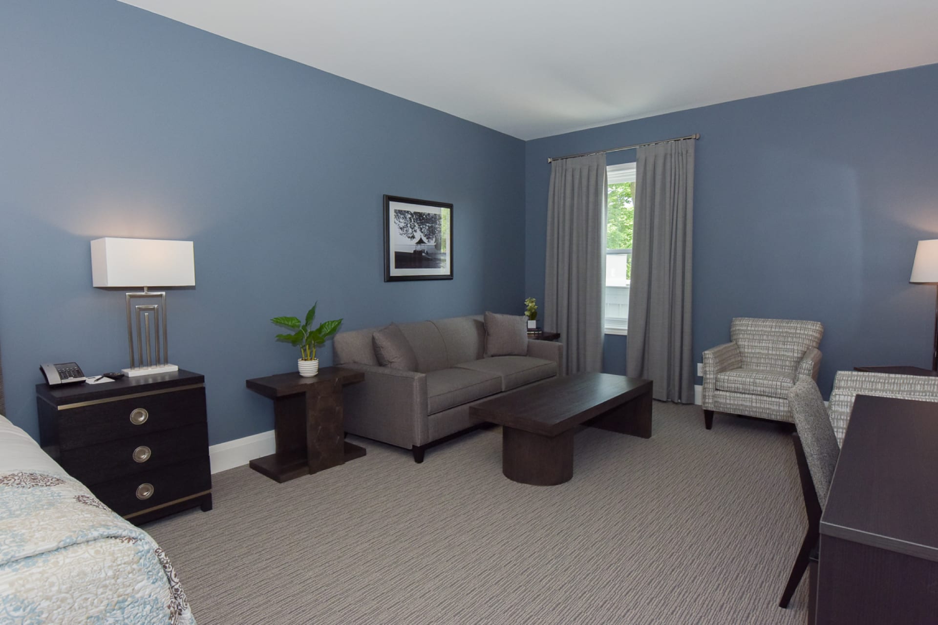 A minimalist living room with blue walls featuring a gray sofa, a wooden coffee table, two patterned armchairs, and a framed black-and-white photo. Two lamps on side tables and a window with gray curtains complete the space. 124 on Queen Hotel and Spa | Hotel & Spa Niagara-on-the-lake | 1 (855) 988-4552