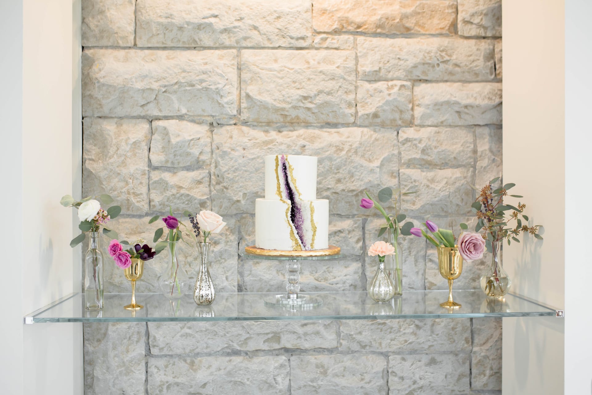 A two-tiered cake with a geode design sits on a glass shelf against a stone wall. The cake is flanked by vases and chalices holding various flowers, including roses and carnations in pink and purple hues. 124 on Queen Hotel and Spa | Hotel & Spa Niagara-on-the-lake | 1 (855) 988-4552