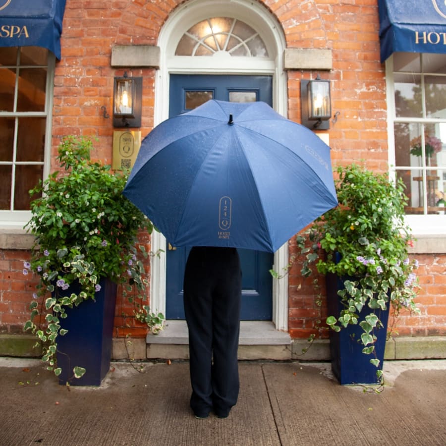 A person stands in front of a brick buildings blue door, holding a large blue umbrella that obscures their upper body. They are flanked by potted plants with cascading greenery and flowers. 124 on Queen Hotel and Spa | Hotel & Spa Niagara-on-the-lake | 1 (855) 988-4552