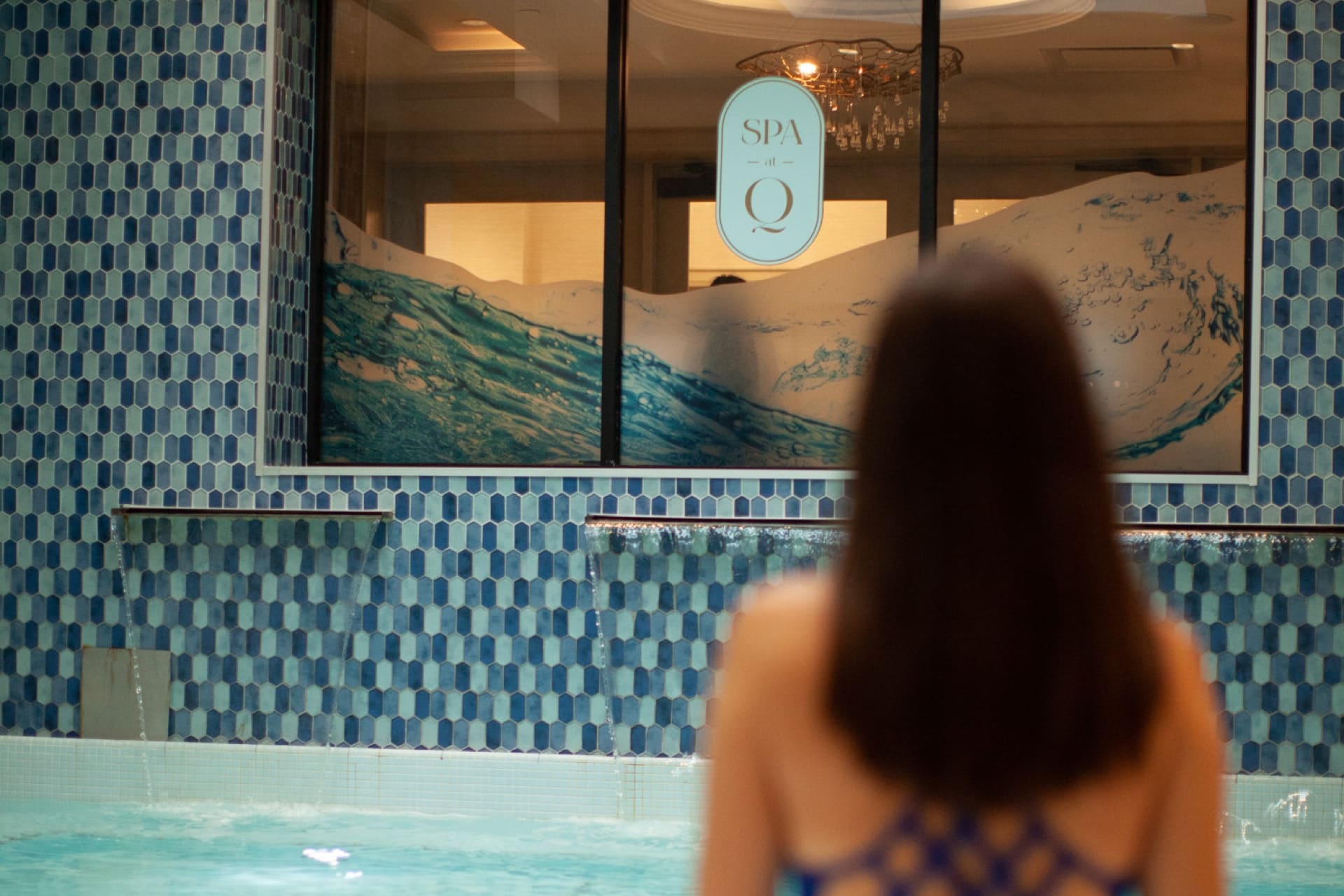A person with long hair, seen from behind, stands by a pool with blue mosaic tiles. A window labeled Spa Q features a wave design. Indoor lighting suggests a relaxing atmosphere. 124 on Queen Hotel and Spa | Hotel & Spa Niagara-on-the-lake | 1 (855) 988-4552