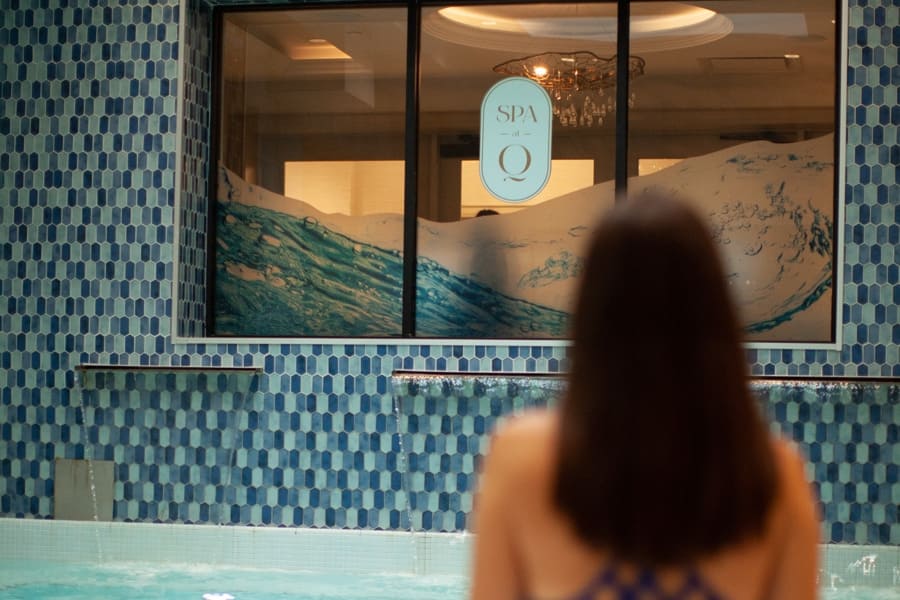 A woman in a blue swimsuit stands at the edge of a pool, facing a window with a spa logo. The window has a wave design, and the surrounding wall is covered in blue mosaic tiles. A chandelier is visible inside. 124 on Queen Hotel and Spa | Hotel & Spa Niagara-on-the-lake | 1 (855) 988-4552