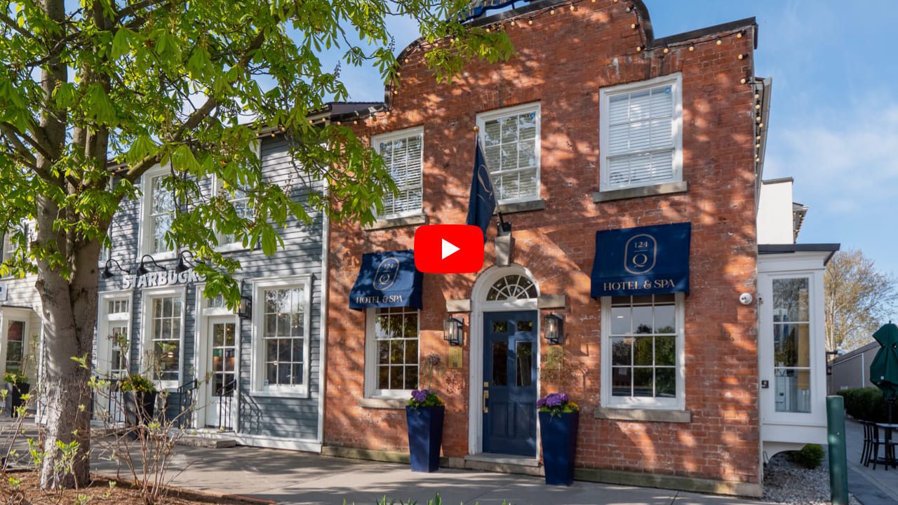 Front view of a charming brick and blue-painted hotel with white-trimmed windows. Blue awnings read Hotel & Spa. A lush tree partially shades the entrance, which is adorned with tall flower pots. A path leads to the entrance. 124 on Queen Hotel and Spa | Hotel & Spa Niagara-on-the-lake | 1 (855) 988-4552