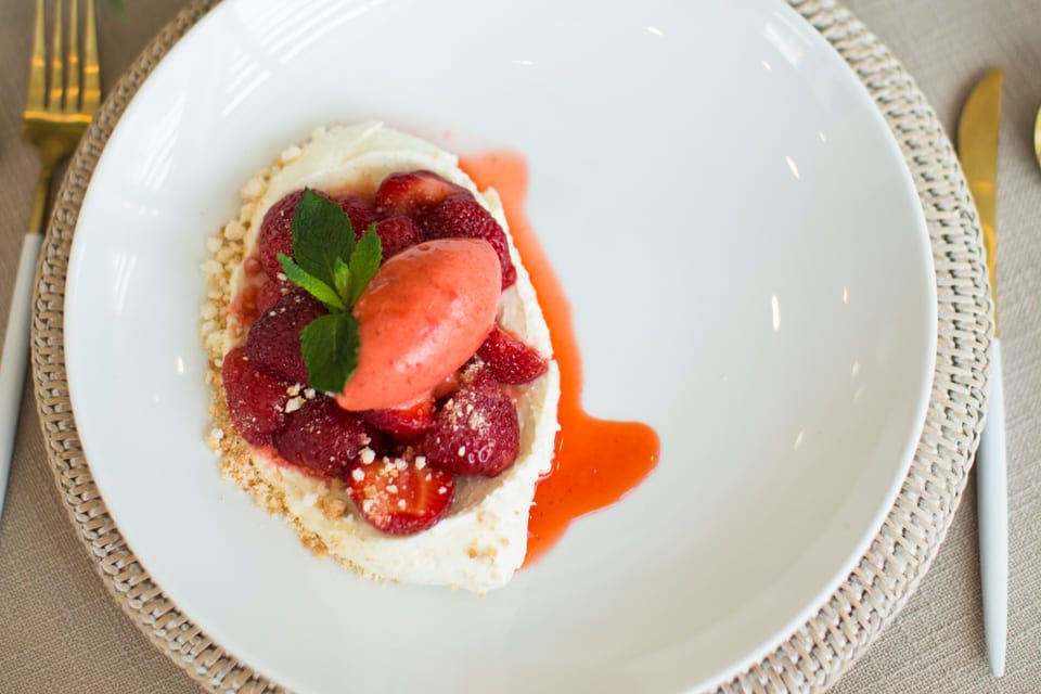 A dessert featuring a white plate with a scoop of pink sorbet on top of sliced strawberries, garnished with a sprig of mint. The dish is served on a woven placemat, with utensils to the side. 124 on Queen Hotel and Spa | Hotel & Spa Niagara-on-the-lake | 1 (855) 988-4552 A dessert featuring a white plate with a scoop of pink sorbet on top of sliced strawberries, garnished with a sprig of mint. The dish is served on a woven placemat, with utensils to the side. 124 on Queen Hotel and Spa | Hotel & Spa Niagara-on-the-lake | 1 (855) 988-4552