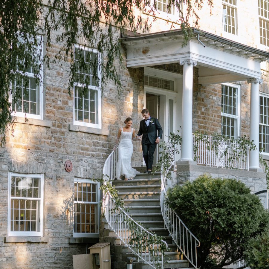 A bride and groom are walking down the steps of a large stone building with white columns and railings adorned with greenery. The bride is in a white dress, and the groom is in a black suit. The scene is set in a lush, green outdoor area. 124 on Queen Hotel and Spa | Hotel & Spa Niagara-on-the-lake | 1 (855) 988-4552 A bride and groom are walking down the steps of a large stone building with white columns and railings adorned with greenery. The bride is in a white dress, and the groom is in a black suit. The scene is set in a lush, green outdoor area. 124 on Queen Hotel and Spa | Hotel & Spa Niagara-on-the-lake | 1 (855) 988-4552