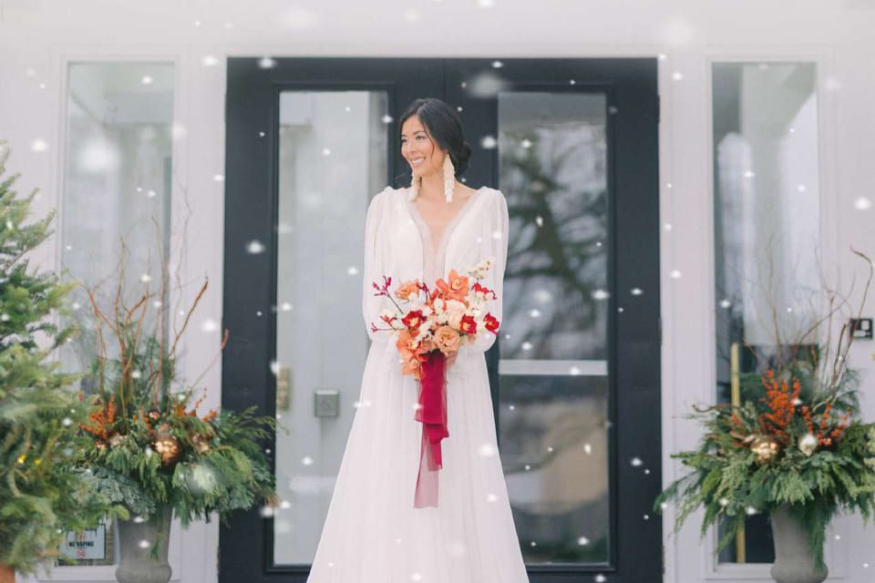 A bride in a white wedding dress stands outside in front of a dark door holding a vibrant bouquet of orange and red flowers. Snowflakes fall gently around her, and festive greenery with branches decorates each side of the entrance. 124 on Queen Hotel and Spa | Hotel & Spa Niagara-on-the-lake | 1 (855) 988-4552 A bride in a white wedding dress stands outside in front of a dark door holding a vibrant bouquet of orange and red flowers. Snowflakes fall gently around her, and festive greenery with branches decorates each side of the entrance. 124 on Queen Hotel and Spa | Hotel & Spa Niagara-on-the-lake | 1 (855) 988-4552