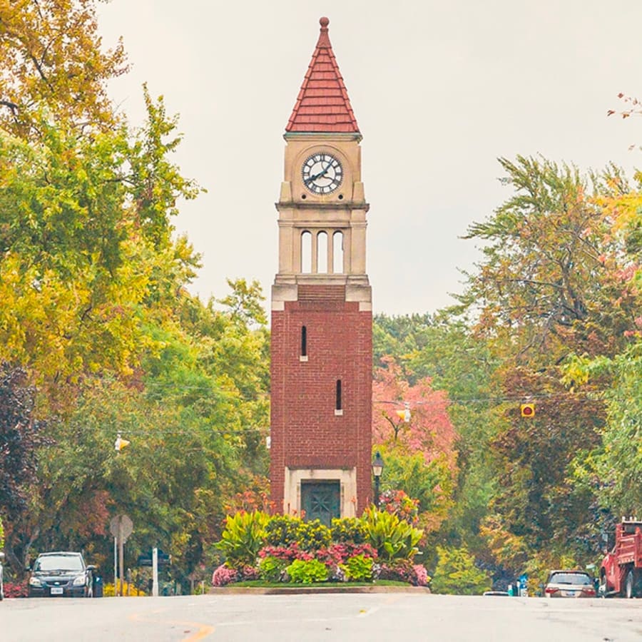 A tall, red-brick clock tower with a pointed roof stands at the center of a tree-lined road. The clock shows the time, and vibrant flowers surround the towers base. Cars are parked along the street, and the trees display autumn colors. 124 on Queen Hotel and Spa | Hotel & Spa Niagara-on-the-lake | 1 (855) 988-4552