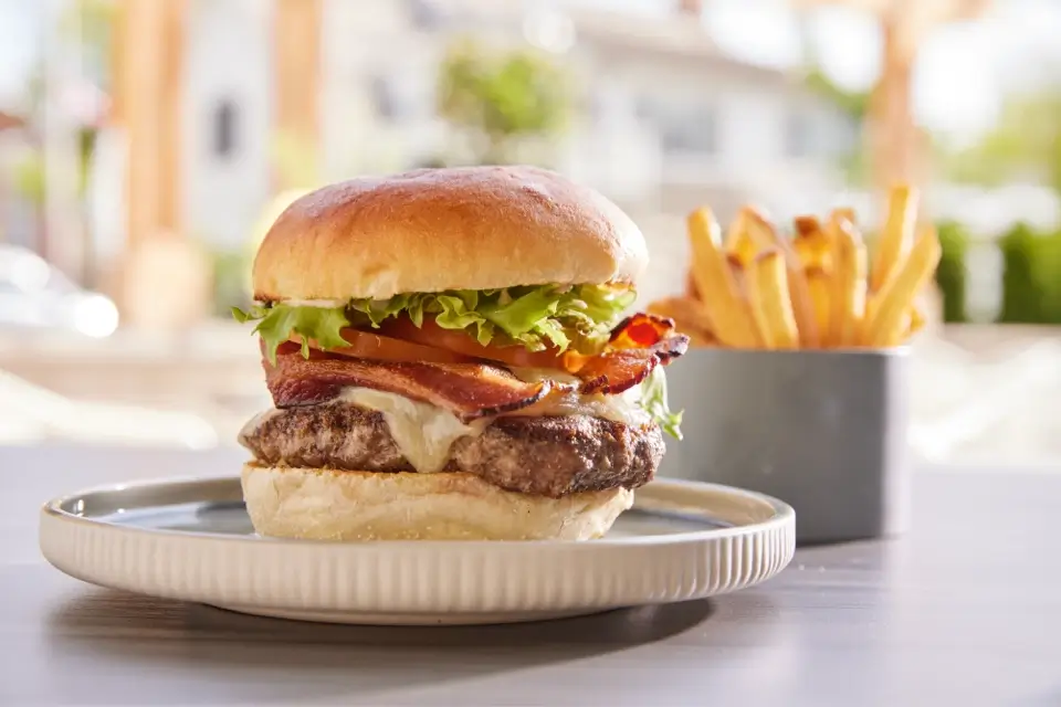 A juicy hamburger on a plate is topped with bacon, lettuce, and cheese, served with a side of crispy French fries in a metal container. The background shows a blurred outdoor setting. 124 on Queen Hotel and Spa | Hotel & Spa Niagara-on-the-lake | 1 (855) 988-4552