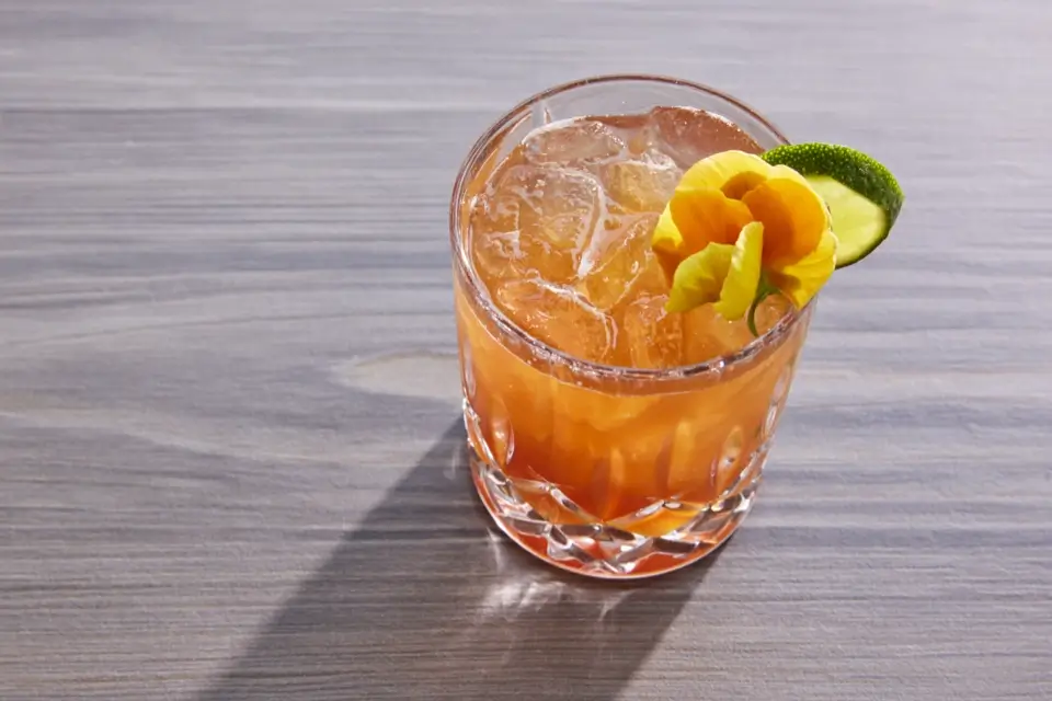 A glass of orange cocktail on a wooden surface, garnished with a yellow flower and lime slice. The drink is filled with ice cubes. 124 on Queen Hotel and Spa | Hotel & Spa Niagara-on-the-lake | 1 (855) 988-4552