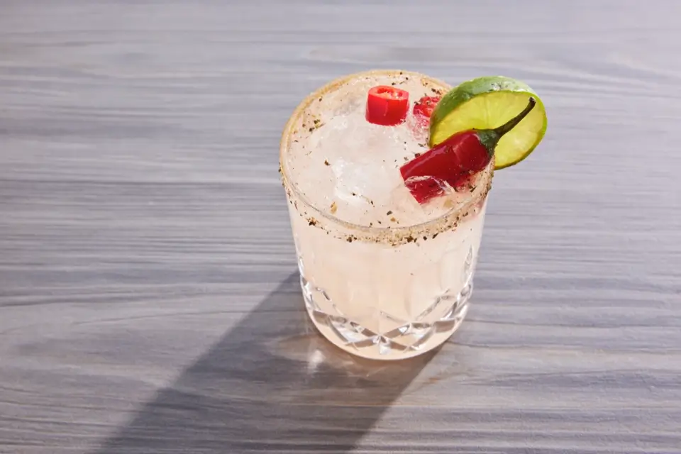 A cocktail in a glass with a salted rim, filled with ice, garnished with a lime slice and a red chili pepper. It is placed on a gray wooden surface. 124 on Queen Hotel and Spa | Hotel & Spa Niagara-on-the-lake | 1 (855) 988-4552