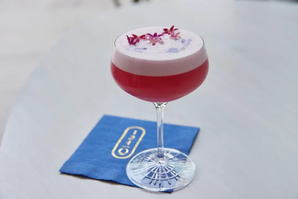 A cocktail in a stemmed glass with a frothy pink top is garnished with small purple and pink flowers. The drink rests on a blue napkin on a light surface. 124 on Queen Hotel and Spa | Hotel & Spa Niagara-on-the-lake | 1 (855) 988-4552