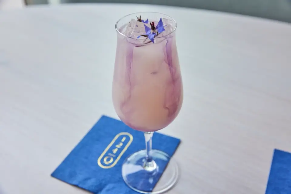 A stemmed glass filled with a lavender-colored drink, garnished with purple flowers, sits on a table. The drink rests on blue napkins with gold lettering. 124 on Queen Hotel and Spa | Hotel & Spa Niagara-on-the-lake | 1 (855) 988-4552
