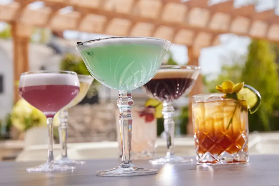 A selection of colorful cocktails served in elegant glassware on an outdoor table. The foreground shows a green drink in a coupe glass. Other drinks include a purple, a dark brown, and an amber cocktail, garnished with lime and flowers. Sunlit pergola above. 124 on Queen Hotel and Spa | Hotel & Spa Niagara-on-the-lake | 1 (855) 988-4552
