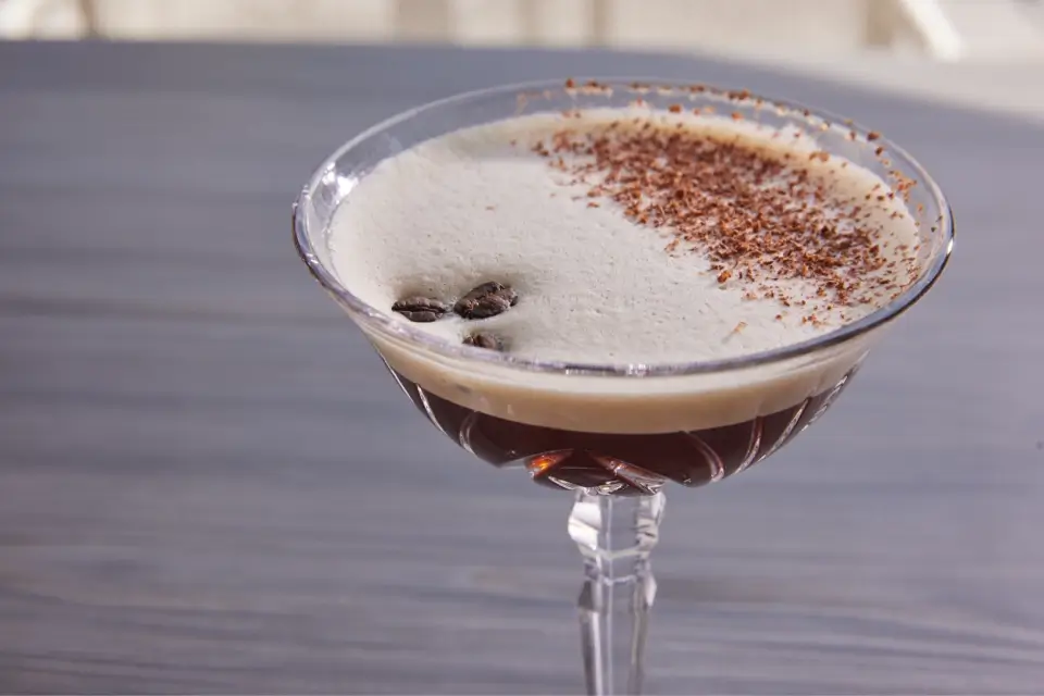 A martini glass filled with an espresso martini cocktail. The drink has a frothy cream layer on top, garnished with three coffee beans and sprinkled with chocolate shavings, set against a gray tabletop background. 124 on Queen Hotel and Spa | Hotel & Spa Niagara-on-the-lake | 1 (855) 988-4552