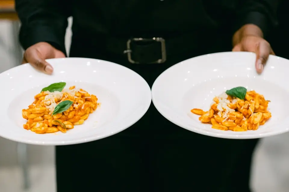 A person holds two white plates, each featuring a serving of pasta with tomato sauce, topped with grated cheese and a basil leaf. The background is blurred, emphasizing the dishes. 124 on Queen Hotel and Spa | Hotel & Spa Niagara-on-the-lake | 1 (855) 988-4552