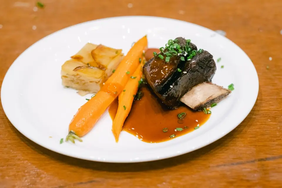 A gourmet dish on a white plate featuring braised short ribs with dark sauce, garnished with chopped herbs. Served alongside are two roasted carrots and a piece of layered potato gratin. The plate rests on a wooden table. 124 on Queen Hotel and Spa | Hotel & Spa Niagara-on-the-lake | 1 (855) 988-4552