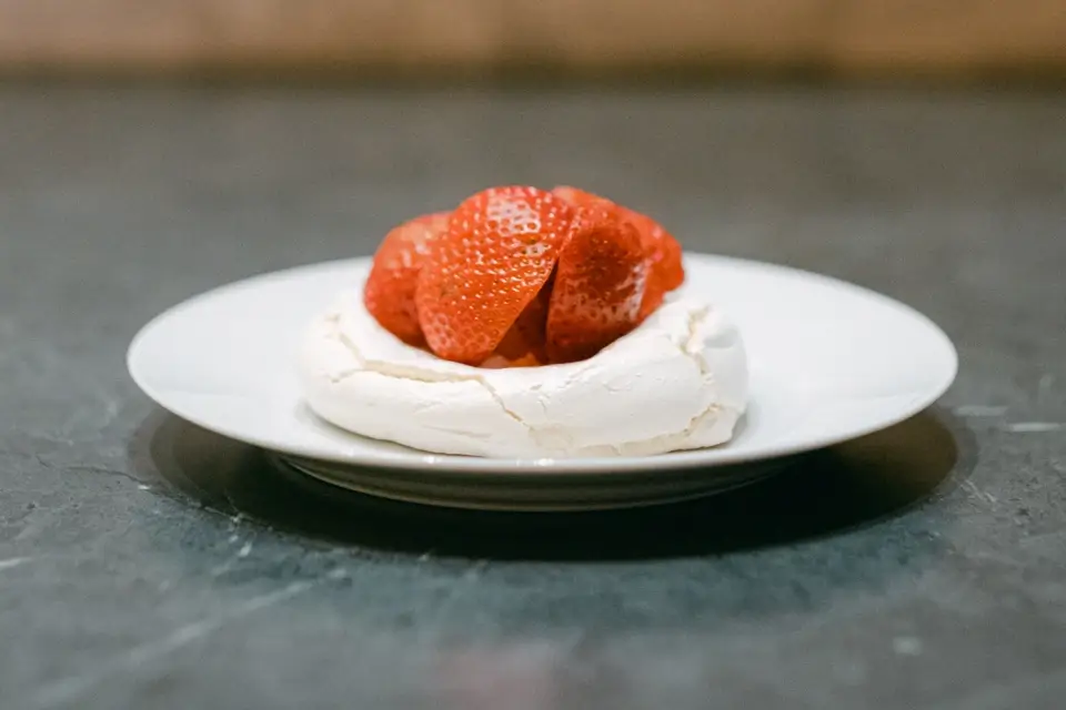 A white meringue dessert topped with sliced strawberries sits on a white plate against a grey background. 124 on Queen Hotel and Spa | Hotel & Spa Niagara-on-the-lake | 1 (855) 988-4552