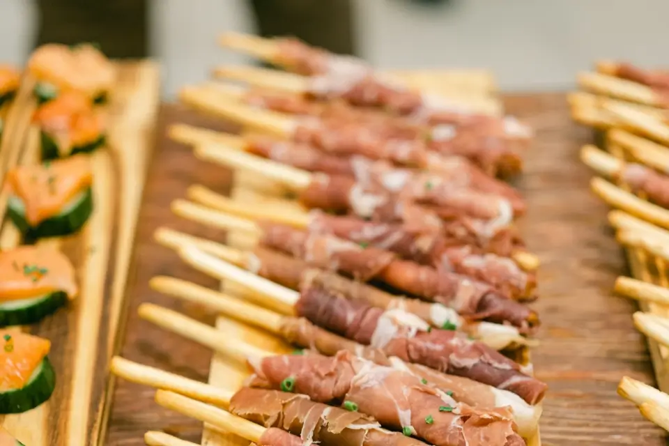 Breadsticks wrapped in prosciutto are neatly arranged on a wooden board. In the background, there are sliced cucumbers topped with a salmon spread on another board. 124 on Queen Hotel and Spa | Hotel & Spa Niagara-on-the-lake | 1 (855) 988-4552