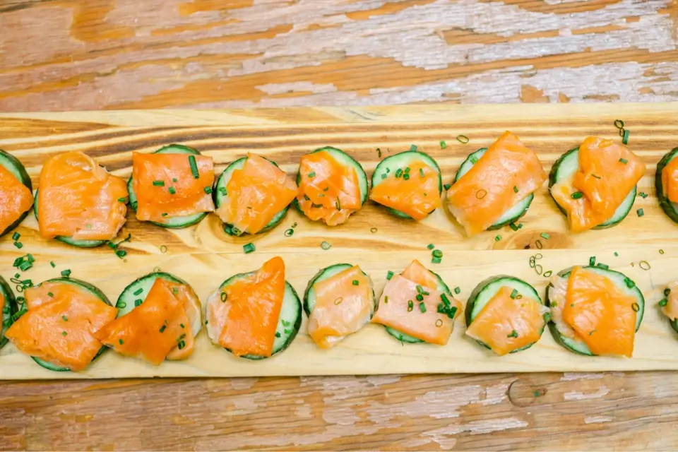 Slices of cucumber topped with smoked salmon and garnished with chopped chives are arranged on a wooden board. The table surface is wooden with a rustic look. 124 on Queen Hotel and Spa | Hotel & Spa Niagara-on-the-lake | 1 (855) 988-4552