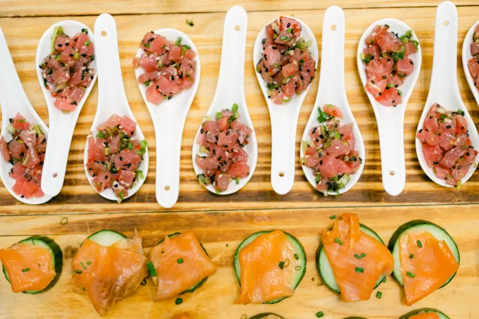 White spoons with diced tuna tartare garnished with sesame seeds and herbs arranged in rows on a wooden surface. Below them, slices of smoked salmon on cucumber rounds, also sprinkled with herbs. 124 on Queen Hotel and Spa | Hotel & Spa Niagara-on-the-lake | 1 (855) 988-4552