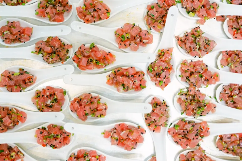 Multiple white ceramic spoons, each holding a small portion of tuna tartare, are arranged on a tray. The tartare is garnished with chopped herbs and black sesame seeds, displaying a fresh, colorful presentation. 124 on Queen Hotel and Spa | Hotel & Spa Niagara-on-the-lake | 1 (855) 988-4552