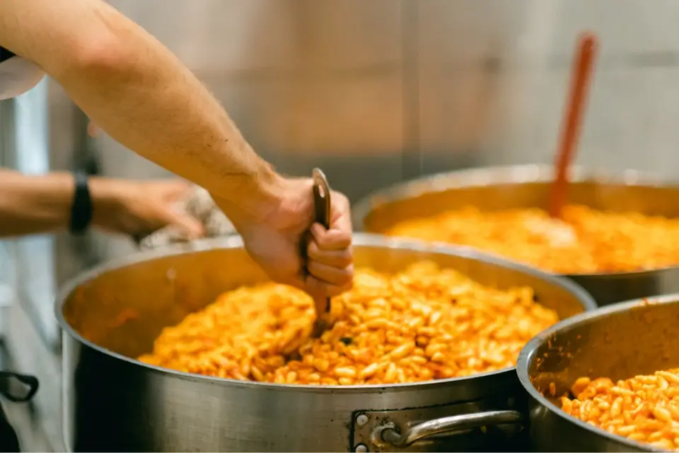 Close-up of a person stirring a large pot filled with cooked beans in a rich, orange sauce. The person uses a wooden spoon and holds a towel in the other hand. Another pot with beans is visible in the background. 124 on Queen Hotel and Spa | Hotel & Spa Niagara-on-the-lake | 1 (855) 988-4552
