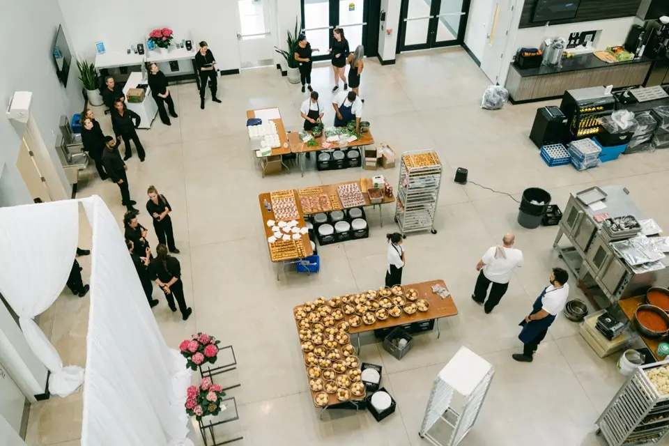 Aerial view of a large kitchen with cooks and staff preparing for an event. Long tables are lined with plates of food, trays, and desserts. Several people are gathered near the entrance, wearing uniforms. Pinks flowers are visible on the bottom left. 124 on Queen Hotel and Spa | Hotel & Spa Niagara-on-the-lake | 1 (855) 988-4552