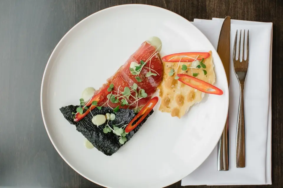 A gourmet dish on a white plate including a piece of rare-cooked meat, black crumbly substance, red pepper slices, and green microgreens. A fork and knife rest on a white napkin beside the plate on a dark wooden table. 124 on Queen Hotel and Spa | Hotel & Spa Niagara-on-the-lake | 1 (855) 988-4552