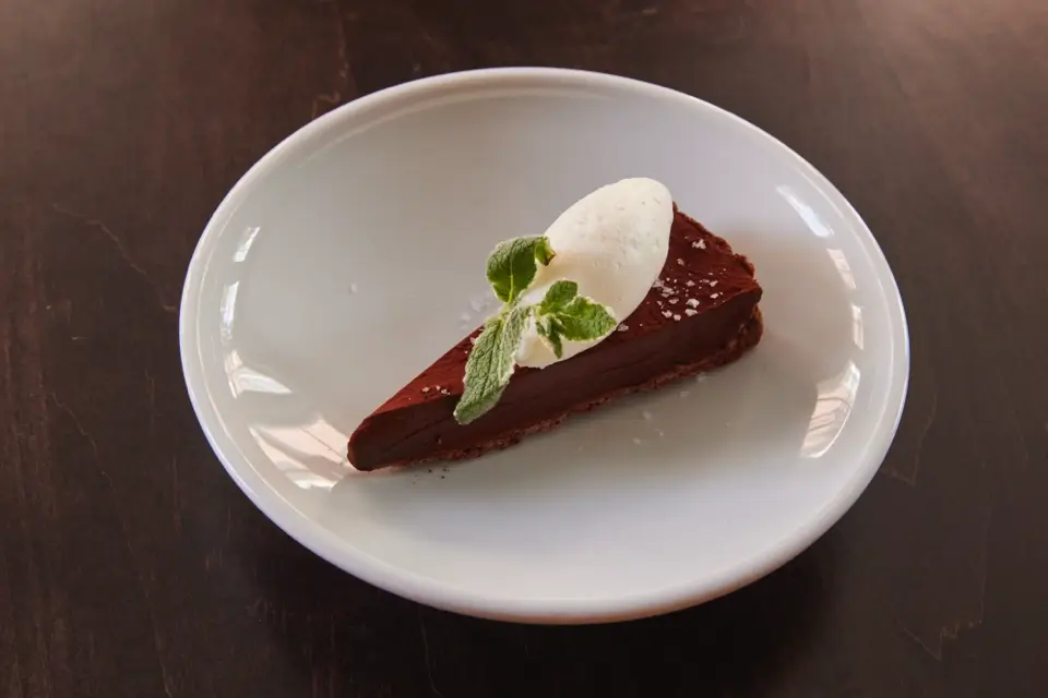 A slice of chocolate cake on a white plate, topped with a dollop of whipped cream and a sprig of fresh mint. The plate is on a dark wooden table. 124 on Queen Hotel and Spa | Hotel & Spa Niagara-on-the-lake | 1 (855) 988-4552