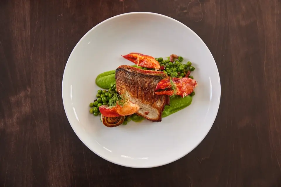 A gourmet dish featuring a fillet of fish with a crispy skin, accompanied by lobster pieces, green pea puree, and whole peas. The meal is artistically plated on a white dish, set against a dark wooden table. 124 on Queen Hotel and Spa | Hotel & Spa Niagara-on-the-lake | 1 (855) 988-4552