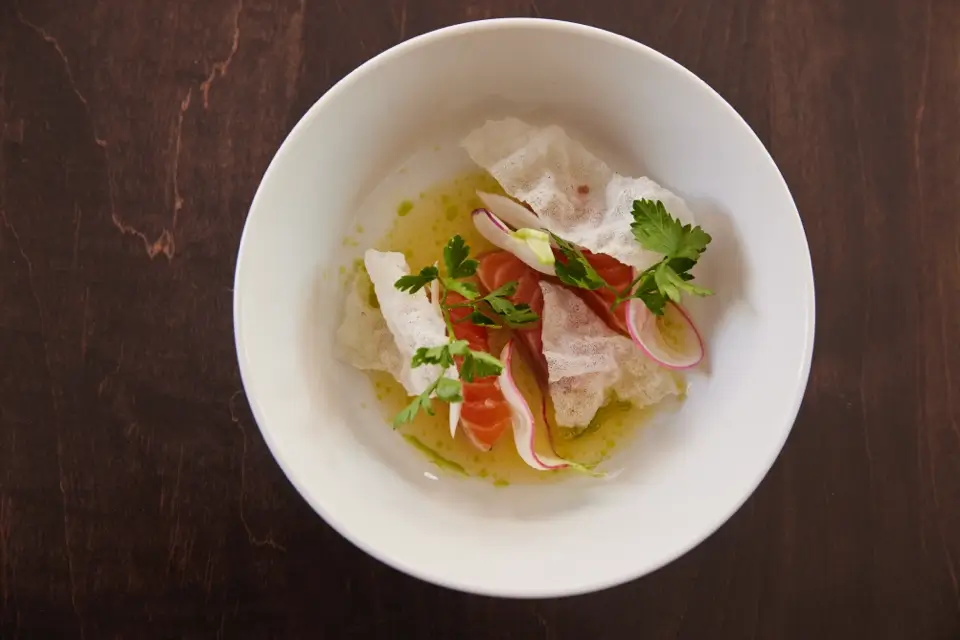 A white bowl on a dark wooden table contains a culinary dish with thinly sliced radishes, salmon, and crispy rice paper on a green sauce. Fresh cilantro leaves serve as garnish. 124 on Queen Hotel and Spa | Hotel & Spa Niagara-on-the-lake | 1 (855) 988-4552