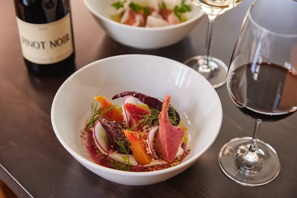 A white bowl containing a gourmet dish with colorful vegetables and herbs, placed on a dark wooden table. Nearby are glasses of red and white wine, along with a bottle labeled Pinot Noir. 124 on Queen Hotel and Spa | Hotel & Spa Niagara-on-the-lake | 1 (855) 988-4552