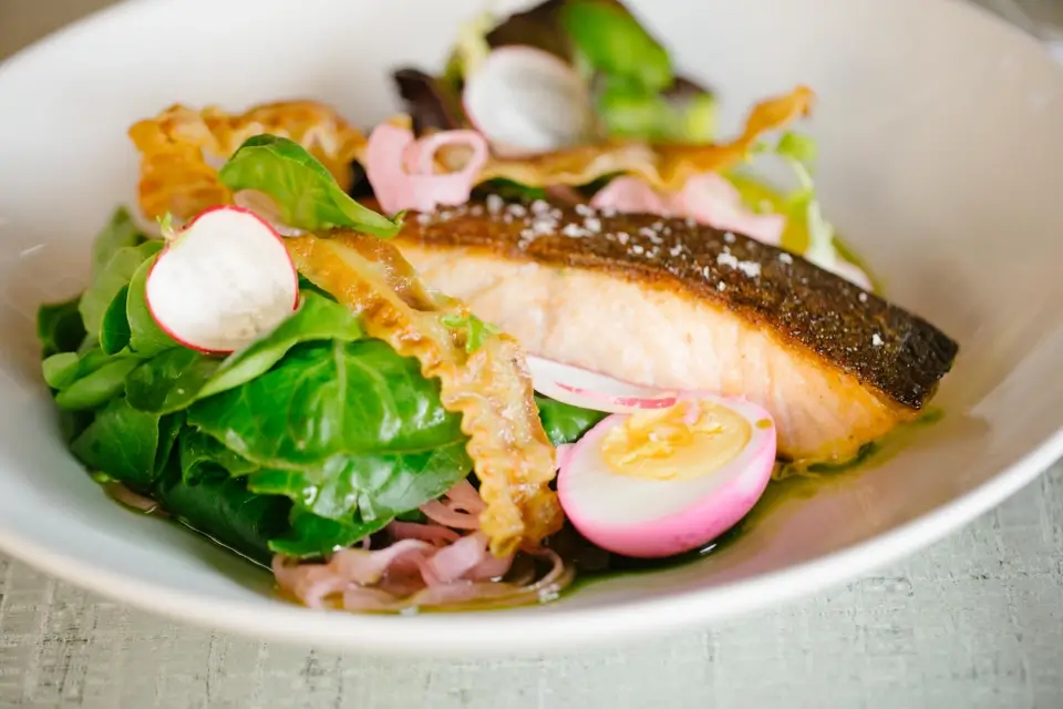 A dish featuring grilled salmon with crispy skin, served atop fresh green salad leaves. The plate includes pink pickled onions, radish slices, and a halved pickled egg, garnished with toasted bread crisps. 124 on Queen Hotel and Spa | Hotel & Spa Niagara-on-the-lake | 1 (855) 988-4552