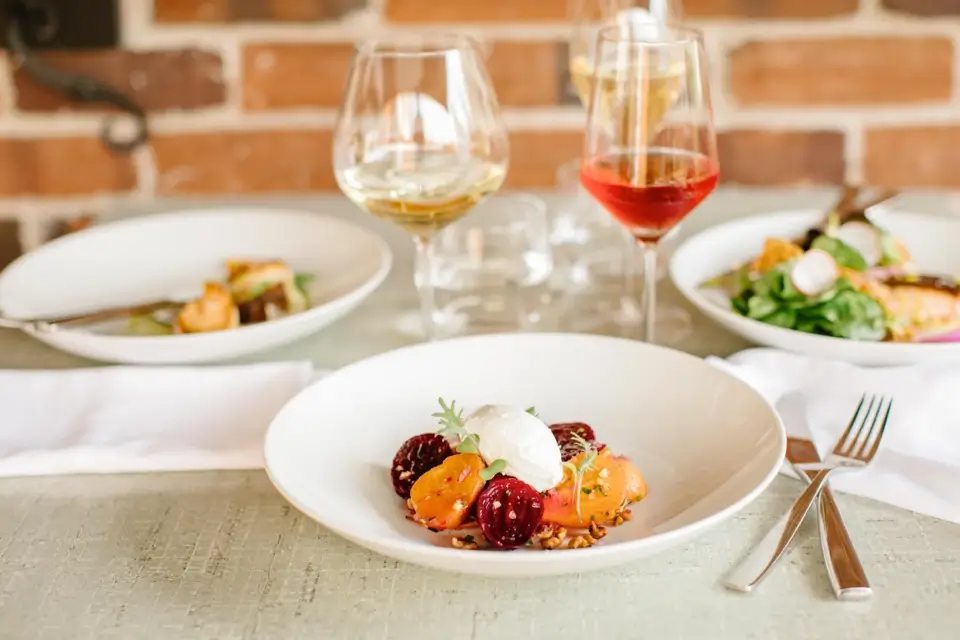 A gourmet dish featuring sliced beets, yellow tomatoes, and a poached egg arranged on a white plate. Two glasses of wine are in the background, with additional plates of food on a rustic light-textured table. 124 on Queen Hotel and Spa | Hotel & Spa Niagara-on-the-lake | 1 (855) 988-4552