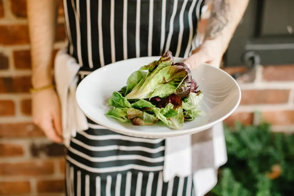A person in a black and white striped apron holds a white plate with a fresh green salad against a brick wall backdrop. They have a towel tucked into their apron pocket. 124 on Queen Hotel and Spa | Hotel & Spa Niagara-on-the-lake | 1 (855) 988-4552