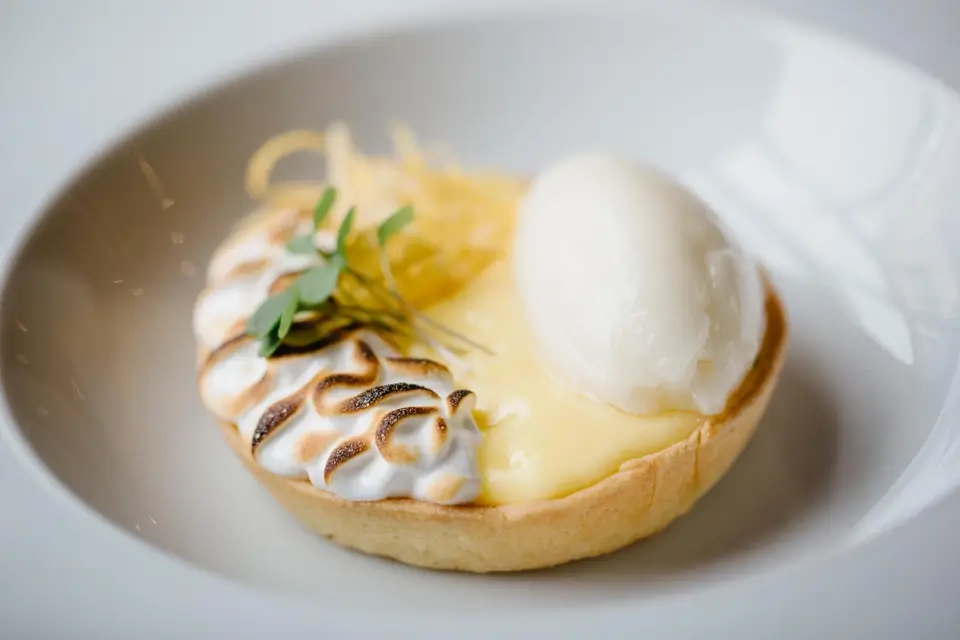 A lemon tart is garnished with toasted meringue, a scoop of sorbet, and a small sprig of green herbs, presented on a white plate. 124 on Queen Hotel and Spa | Hotel & Spa Niagara-on-the-lake | 1 (855) 988-4552