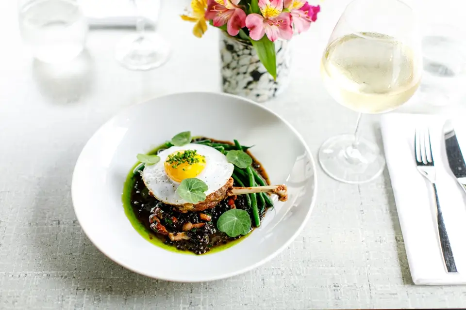 A white plate with gourmet food: a poached egg on a bed of grains and vegetables, garnished with greens. Theres a glass of white wine, a bouquet of flowers, and neatly set cutlery on a textured white tablecloth. 124 on Queen Hotel and Spa | Hotel & Spa Niagara-on-the-lake | 1 (855) 988-4552