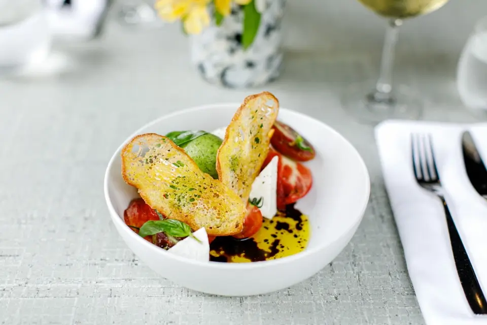 A white bowl containing tomato slices, mozzarella, basil, two pieces of toasted bread, olive oil, and balsamic vinaigrette. Its set on a table with cutlery, a napkin, and a glass of white wine in the background. 124 on Queen Hotel and Spa | Hotel & Spa Niagara-on-the-lake | 1 (855) 988-4552