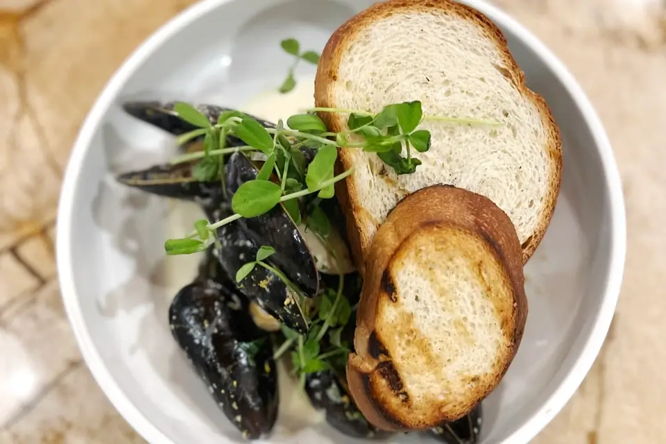 A white bowl contains mussels in a creamy sauce, garnished with green microgreens. Two slices of toasted bread rest on top, slightly overlapping each other. The bowl sits on a marble countertop. 124 on Queen Hotel and Spa | Hotel & Spa Niagara-on-the-lake | 1 (855) 988-4552