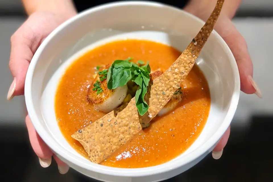 A person holds a white bowl containing tomato soup garnished with a seared scallop, fresh basil leaves, and a long, crispy cracker. 124 on Queen Hotel and Spa | Hotel & Spa Niagara-on-the-lake | 1 (855) 988-4552