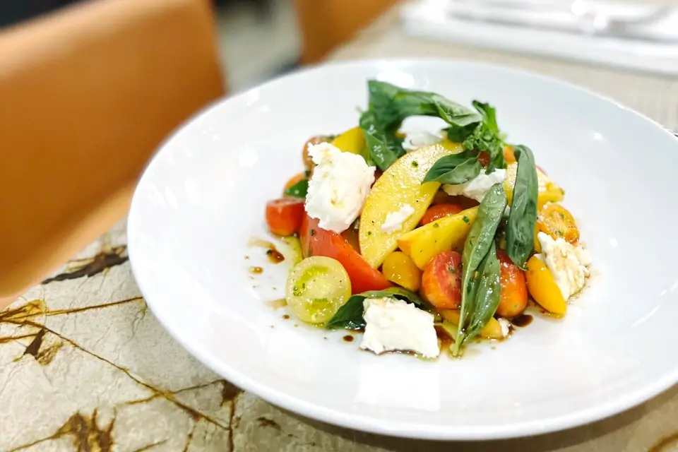 A white plate with a colorful salad featuring sliced heirloom tomatoes, mozzarella cheese, fresh basil leaves, and a drizzle of balsamic vinegar. The plate is set on a marble-patterned table. 124 on Queen Hotel and Spa | Hotel & Spa Niagara-on-the-lake | 1 (855) 988-4552