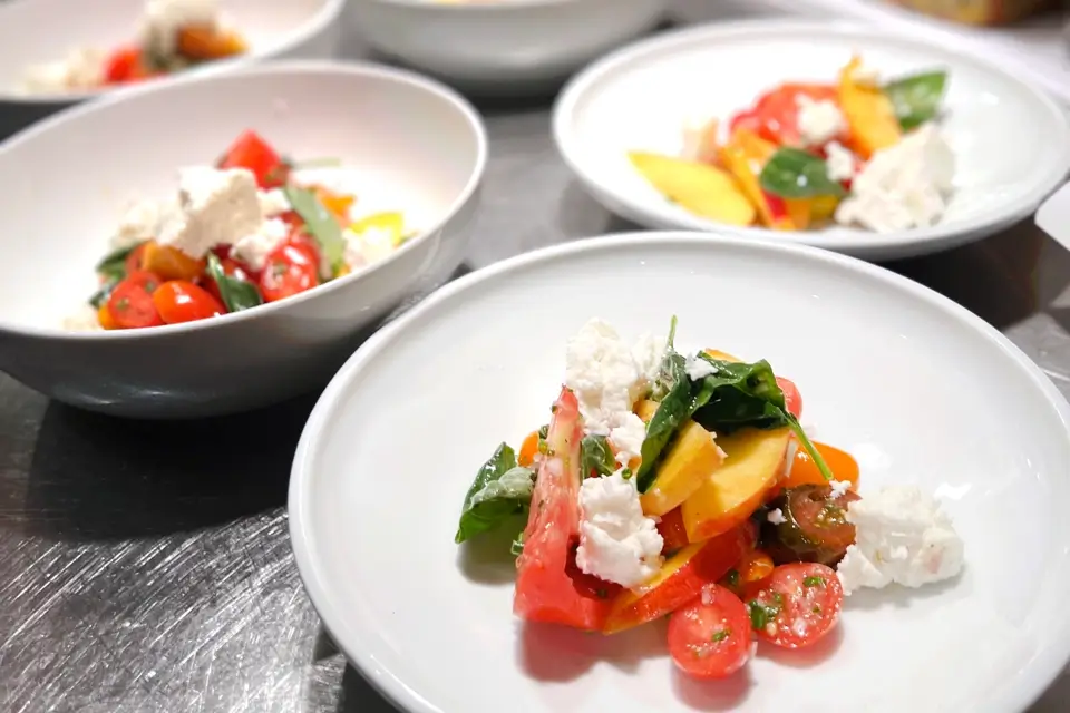 Three white bowls filled with a colorful salad featuring sliced heirloom tomatoes, fresh greens, crumbled cheese, and herbs, arranged on a stainless steel surface. The vibrant ingredients create a visually appealing, fresh dish. 124 on Queen Hotel and Spa | Hotel & Spa Niagara-on-the-lake | 1 (855) 988-4552