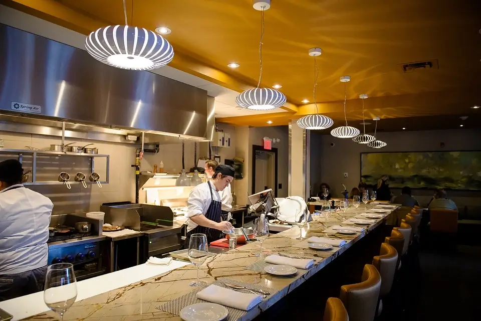 A modern restaurant kitchen with chefs preparing food. The long counter is set with plates, cutlery, and wine glasses. Contemporary pendant lights hang from the ceiling, casting warm light over the scene. A large landscape painting is on the wall. 124 on Queen Hotel and Spa | Hotel & Spa Niagara-on-the-lake | 1 (855) 988-4552