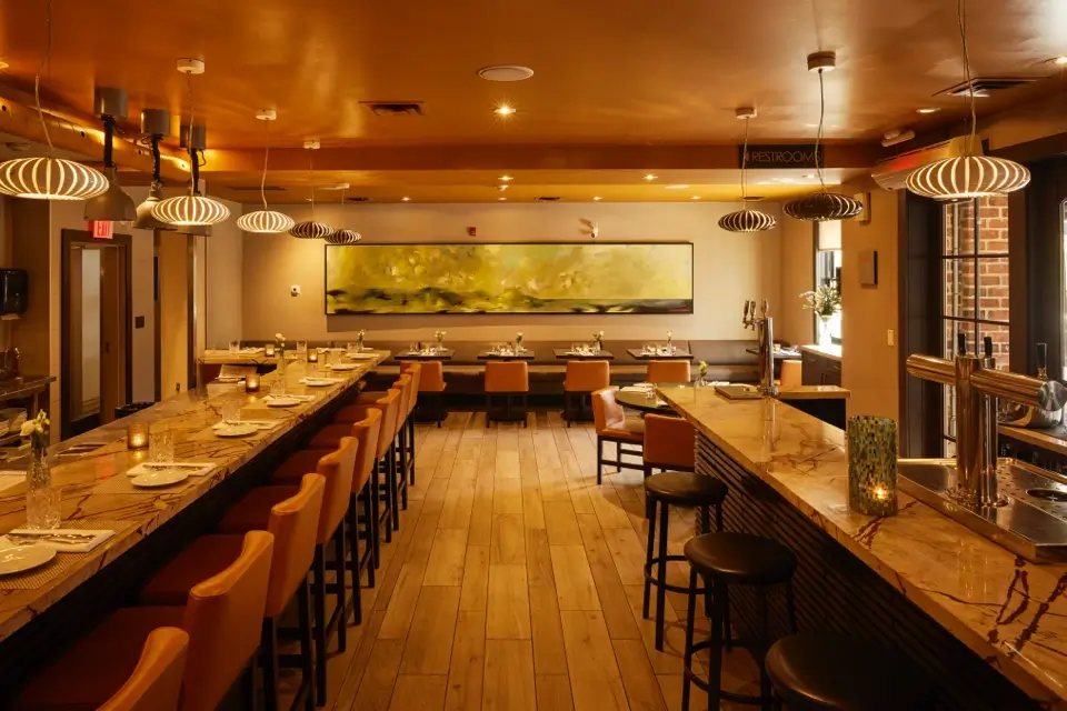 A warmly lit restaurant interior with wood flooring, featuring a long bar with stools on the left and dining tables with chairs on the right. Modern pendant lights hang from the ceiling, and artwork decorates the far wall. 124 on Queen Hotel and Spa | Hotel & Spa Niagara-on-the-lake | 1 (855) 988-4552
