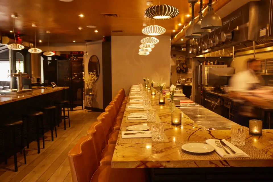 A warmly lit restaurant interior featuring a long, marble-topped counter with leather chairs on one side. It has neatly set plates, cutlery, and glasses. The open kitchen is visible in the background, and decorative lights hang from the ceiling. 124 on Queen Hotel and Spa | Hotel & Spa Niagara-on-the-lake | 1 (855) 988-4552
